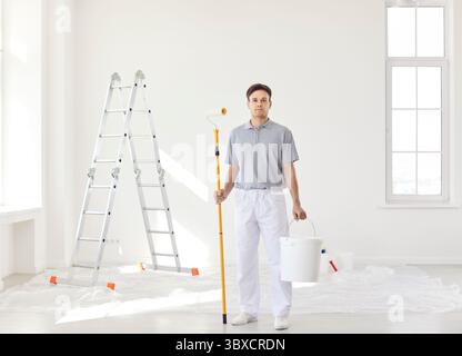Peintre professionnel confiant avec rouleau de peinture et seau dans les mains regardant la caméra Banque D'Images