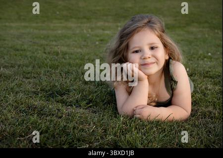 Fille enfant souriante couchée sur l'herbe, regardant la caméra. Menton sur les mains. Banque D'Images