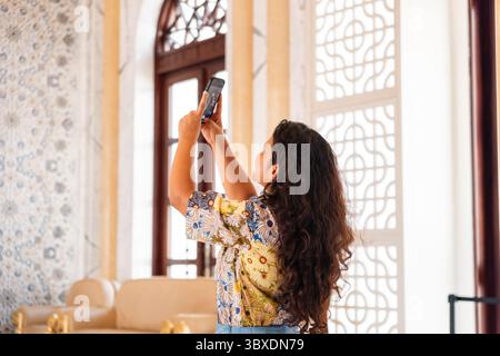 Une femme prenant une photo avec un smartphone, à l'intérieur d'une pièce élégante avec des murs à motifs. Elle porte une chemise fleurie et a de longs cheveux bouclés Banque D'Images