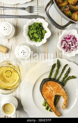 Steak de saumon sur un lit d'asperges, accompagné d'herbes fraîches et d'oignons. Un verre de vin blanc et un bol de pommes de terre complètent l'alléchant ensemble de repas Banque D'Images