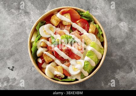 Bol en papier jetable avec salade César appétissante de laitue romaine, tomates cerises, œufs de caille, croûtons et filet de saumon fumé assaisonné de pi Banque D'Images