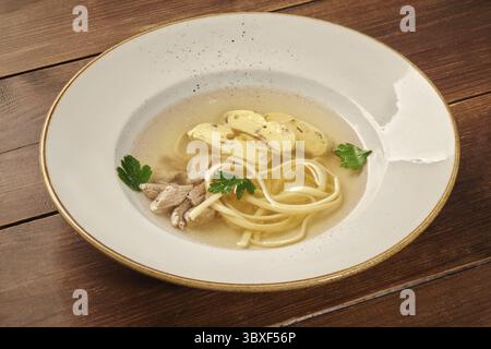 Soupe chaude et réconfortante de consommé de poulet avec nouilles aux œufs, omelette tranchée, persil frais, servie dans un bol blanc sur une table en bois rustique Banque D'Images