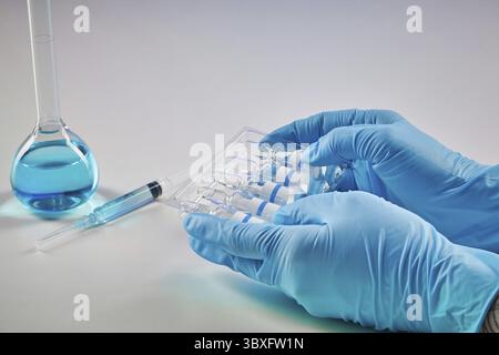 Mains dans des gants jetables contenant un jeu de cinq ampoules de liquide. Flacon médical avec réactif chimique bleu, seringue isolée sur blanc. Laboratoire r Banque D'Images