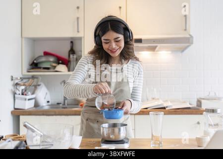 Une femme portant des écouteurs verse des ingrédients dans un bol, souriant alors qu'elle aime la musique avec sa cuisson dans une cuisine familiale Banque D'Images