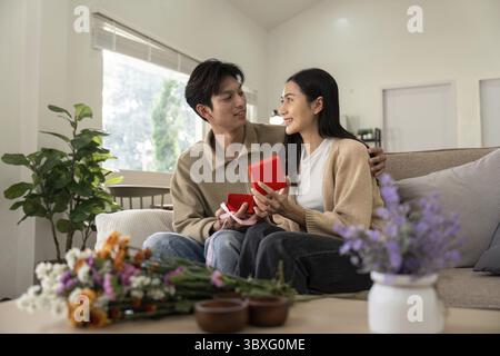 Un couple joyeux assis sur un canapé, échangeant des cadeaux dans un salon confortable rempli de fleurs et de lumière naturelle Banque D'Images