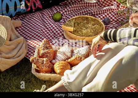 Scène de pique-nique d'été confortable le jour ensoleillé sur l'herbe verte avec des croissants moelleux doux dans un panier en osier, quiche salée maison et fruits frais dispersés Banque D'Images
