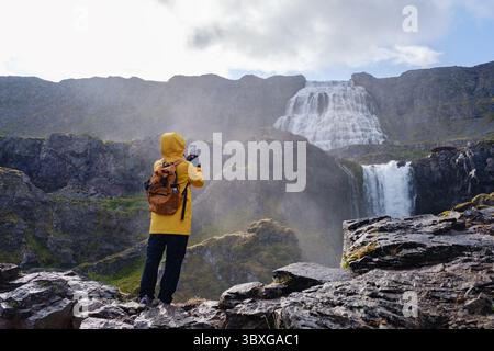 Un voyageur vêtu d'une veste de pluie jaune se tient sur un terrain rocheux, capturant la beauté de la cascade de Dynjandi, Westfjord, Islande. La cascade se déverse dramatiquement dans un paysage luxuriant sous un ciel nuageux Banque D'Images