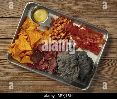 Plateau métallique avec chips de tortilla au maïs croustillantes, arachides frites glacées salées épicées, diverses viandes séchées et sauce au fromage dans un petit bol sur fond de bois Banque D'Images