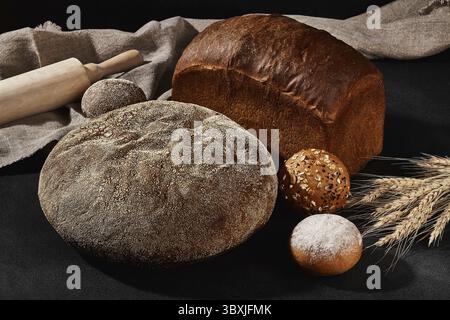 Variété de délicieux pains et pains cuits au four avec graines de sésame et de tournesol, saupoudrés de farine. Bouquet de blé, burlap, goupille de roulement, fond noir, co Banque D'Images