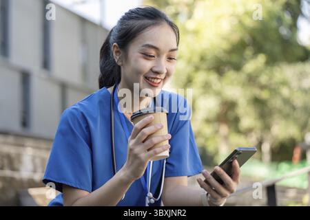 Une travailleuse de santé asiatique en gommage bleu sourit tout en dégustant un café et en utilisant son smartphone pendant une pause à l'extérieur Banque D'Images