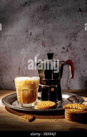 Café chaud servi dans un verre avec de la mousse, à côté d'un pot de moka vintage et des biscuits sur un plateau en métal, posé sur une table en bois avec un fond rustique. Banque D'Images