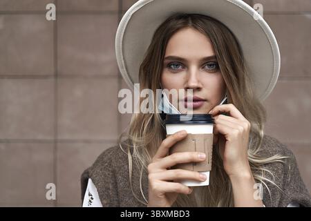 Fin du verrouillage de quarantaine. Une fille vaccinée boit du café tout en tirant sur le masque de protection Banque D'Images