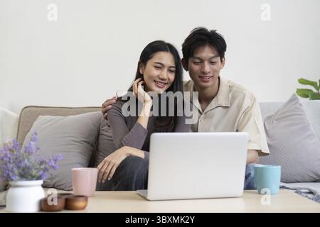 Un jeune couple s'assoit sur le canapé, souriant alors qu'ils partagent un moment agréable tout en regardant du contenu sur un ordinateur portable Banque D'Images