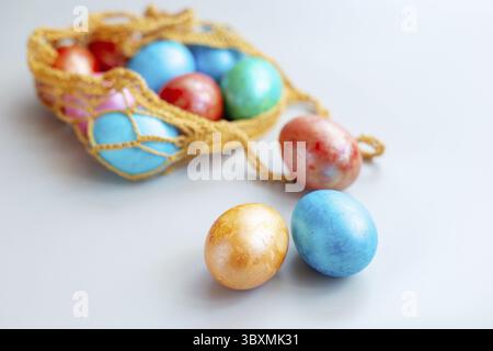 Plusieurs oeufs multicolores se trouvent dans un sac tricoté sur une table blanche. Trois œufs reposent à proximité sur un fond blanc. Copier l'espace Banque D'Images