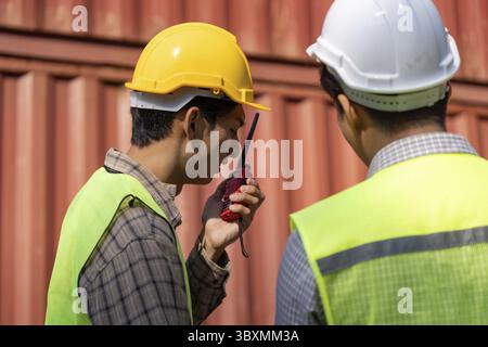 Deux ingénieurs en construction travaillant sur les équipements de sécurité examinent les plans tout en discutant de la logistique à l'aide d'un talkie-walkie Banque D'Images