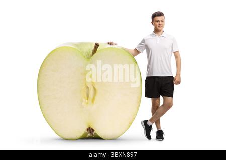 Portrait plein pied d'un jeune homme en sportswear appuyé sur une grosse pomme coupée en deux isolé sur fond blanc Banque D'Images