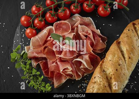 Délicieux, tranchés manuellement jamon, tomates cerises, baguette, fines herbes et épices sur ardoise noir bord contre un fond gris foncé. La cuisine, reste Banque D'Images