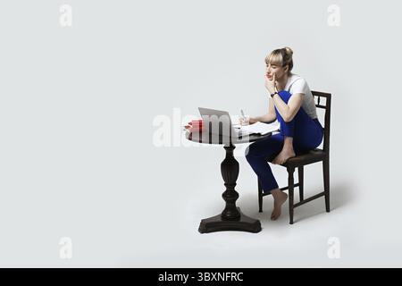 Jeune étudiante concentrée assise au bureau travaillant ou étudiant à distance en ligne écrivant des notes tout en regardant sur l'écran d'un ordinateur portable. Apprentissage à distance ou rem Banque D'Images