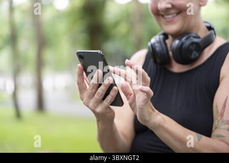 Gros plan des mains d'une femme âgée à l'aide d'un smartphone, avec des écouteurs autour du cou, mettant en évidence l'utilisation de la technologie dans un cadre naturel Banque D'Images