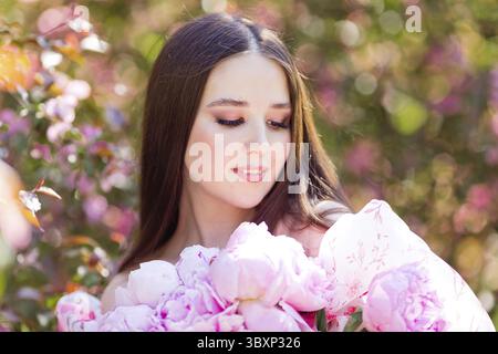Gros plan d'une belle fille souriante avec un grand bouquet de pivoines roses, se tient dans un jardin par une journée ensoleillée. Copier l'espace Banque D'Images