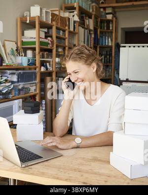 Fabrication de produits, pièce à usiner. Jeune femme entrepreneur designer parlant sur le téléphone portable et regardant l'ordinateur portable, dame souriante dans l'atelier. WOM Banque D'Images