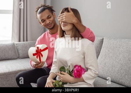 Un gars dans un pull rose et une fille dans une robe blanche, sur un canapé gris . Le gars a couvert les yeux des filles avec sa main pour donner une surprise - un coeur blanc- Banque D'Images