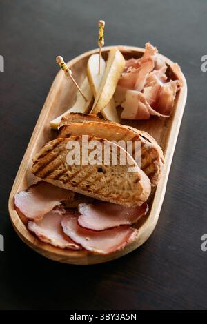 Un plateau de charcuterie composé de pain grillé, de prosciutto, de poires tranchées et de viandes fumées présentées sur un plateau en bois, parfait pour les repas gastronomiques Banque D'Images