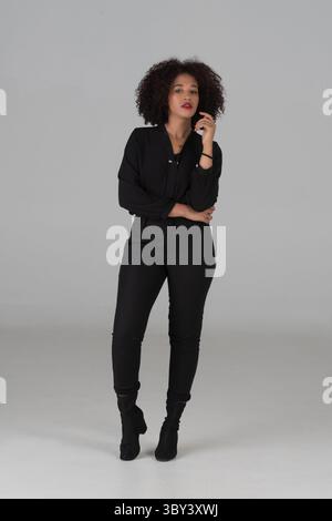 Une jeune femme en tenue noire chic. Femme à la mode avec les cheveux bouclés et rouge à lèvres frappe une pose dans un studio élégant de haut noir et de pantalon sur gra Banque D'Images