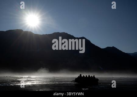 Scène rétro-éclairée du coucher de soleil sur des montagnes sombres reflétant sur le brouillard et les passagers dans un Zodiac depuis un navire de croisière d'expédition dans le Vikingebugt, A. Banque D'Images