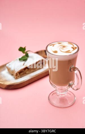 Un cappuccino magnifiquement présenté avec latte art est assis à côté d'une tranche de strudel de pomme, garnie de menthe, sur un fond rose. Banque D'Images