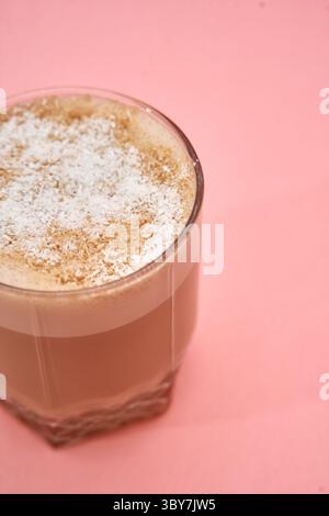 Un chocolat chaud crémeux à la noix de coco est élégamment servi dans un verre sur un fond rose tendre, surmonté de poudre de noix de coco saupoudrée. Banque D'Images