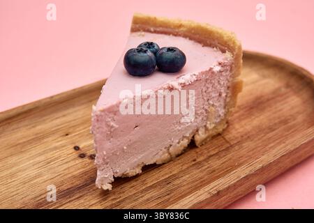 Une tranche de cheesecake rose garnie de myrtilles repose élégamment sur une assiette en bois sur un fond rose pastel, offrant une délicieuse et ind Banque D'Images