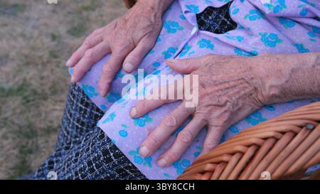 Mains âgées reposant sur la robe florale dans une chaise en osier Banque D'Images