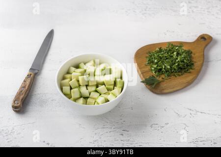 Nourriture, Un bol de courgettes coupées en dés, une planche de bois avec des herbes et un couteau sur un fond bleu clair. Cuisine délicieux petit déjeuner maison Banque D'Images