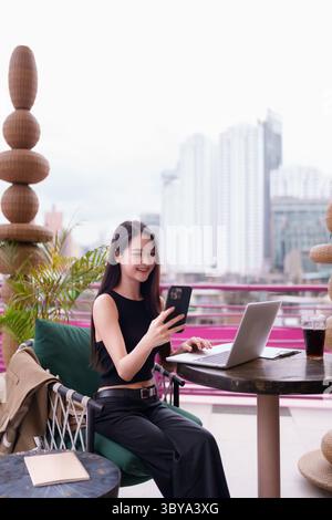Jeune femme asiatique adulte confiante travaille à distance dans un café en plein air. Elle utilise un ordinateur portable et un téléphone pour les tâches professionnelles bénéficiant d'une vie moderne et flexible Banque D'Images