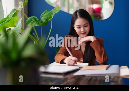 Professionnel asiatique confiant prenant des notes avec diligence à son bureau dans un environnement de bureau élégant. Empowered Woman s'est concentrée sur son travail moderne. Banque D'Images