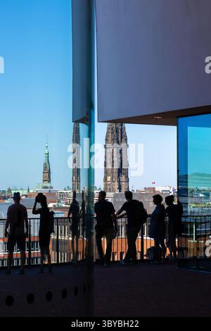 Hambourg, Allemagne - 7 juillet 2025 : silhouette des gens sur un balcon d'Elbphilharmonie avec une vue sur le paysage urbain de Hambourg, y compris les tours et le ciel bleu. Banque D'Images