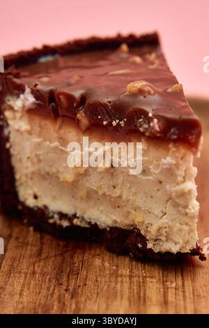 Une délicieuse tranche de cheesecake au beurre de cacahuète au chocolat garnie d'une riche ganache, présentée sur une planche de bois sur un fond rose. Banque D'Images
