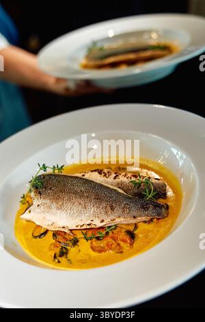 Bar grillé servi avec une sauce à base de safran et garni d'herbes fraîches, élégamment étalé sur des plats blancs, mettant en valeur la gastronomie gastronomique Banque D'Images