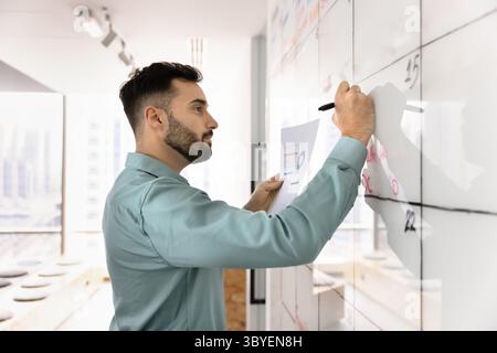 Homme sérieux tenant des papiers avec des graphiques et écrivant sur le tableau blanc Banque D'Images