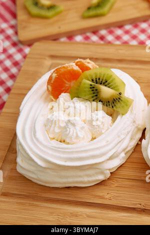 Un gros plan d'un dessert pavlova orné de crème fouettée, de tranches de mandarine et de kiwi sur une planche de bois, mettant en valeur un mélange parfait de moelleux, sucré, Banque D'Images