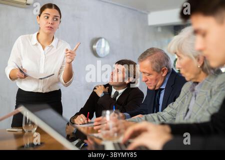 Intelligent jeune femme faisant rapport tandis que les employés de bureau d'âges différents utilisant des cahiers à la table dans le bureau Banque D'Images
