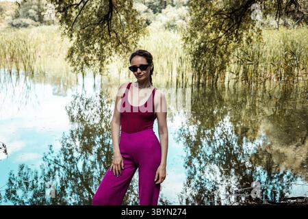 Belle femme en tenue violette pose près du lac tranquille en été. Une femme portant des lunettes de soleil noires pose devant un étang pittoresque ou une rivière, dr. Banque D'Images