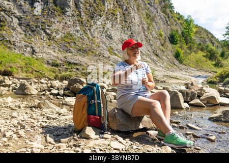 Une femme âgée portant une casquette rouge et une chemise rayée est assise sur un rocher dans un ruisseau de montagne rocheux, profitant d'un verre d'eau pendant une pause randonnée. Son backpa Banque D'Images