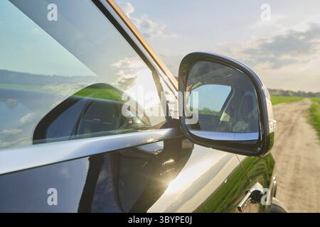Un paysage vibrant se reflète dans le rétroviseur latéral d'une voiture garée. Le soleil brille brillamment sur le véhicule tandis que les champs verts s'étendent dans le distan Banque D'Images