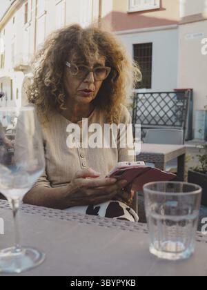 Deux femmes caucasiennes âgées avec les cheveux courts et bouclés respectivement sont assis à une table d'un café en plein air dans une journée d'été, en profitant de boissons et de conv Banque D'Images