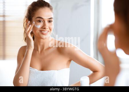 Jeune femme appliquant de la crème sur les joues regardant Reflection Indoor Banque D'Images