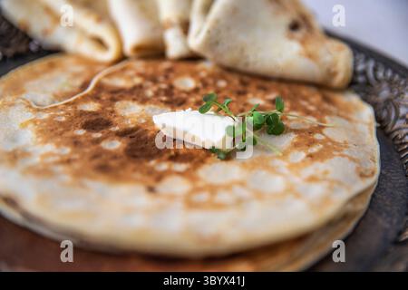 Gros plan d’une crêpe pliée garnie d’une cuillère de fromage à la crème et de microverts frais sur une assiette ornée, évoquant un brunch élégant et un charme artisanal. Banque D'Images