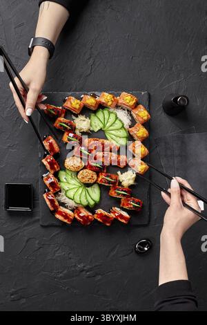 Deux mains femelles utilisant des baguettes pour prendre des rouleaux d'un ensemble de délicieux uramaki de saumon, sushi nigiri et gunkanmaki servis sur une planche d'ardoise avec déchiqueté Banque D'Images