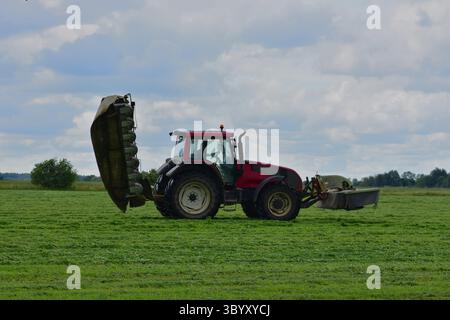 Vue latérale d'un tracteur rouge avec équipement de fauche relevé sur un terrain herbeux sous un ciel nuageux. Machines agricoles pendant le travail saisonnier. Banque D'Images